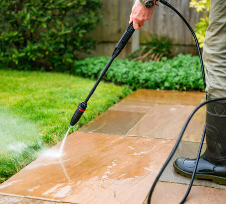 h3 A senior man in his 70s cleans his patio stone slabs using a pressure washer. The man wears a green and blue check shirt with a green gilet.